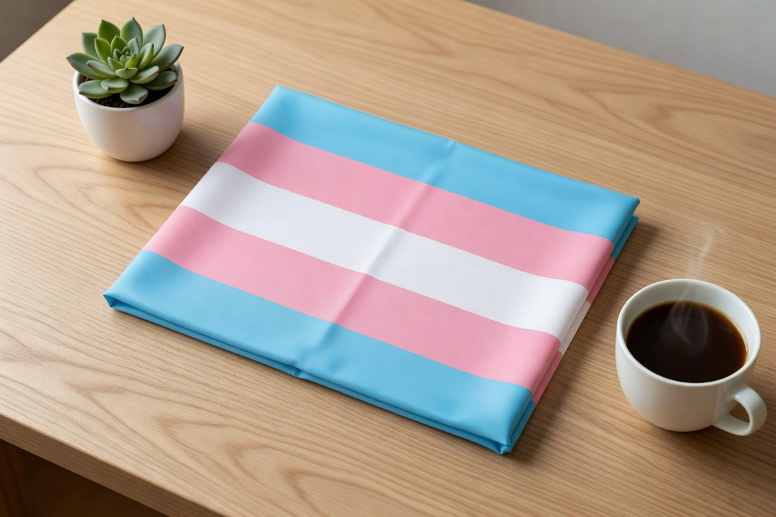 Folded transgender pride flag with blue, pink, and white stripes on a light oak table with a succulent and coffee cup
