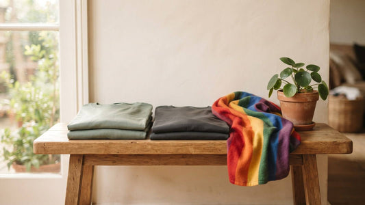 Folded sage and charcoal shirts beside rainbow fabric and a plant on a sunlit wooden table