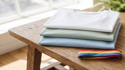 Folded pastel cotton T-shirts and a rainbow fabric strip on a wood bench in window light