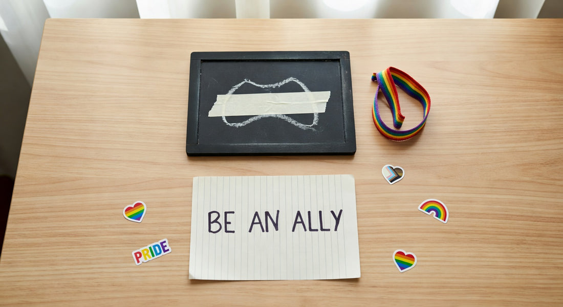 Flat lay with chalkboard showing taped mouth, BE AN ALLY note, rainbow wristband, and pride stickers on wooden desk