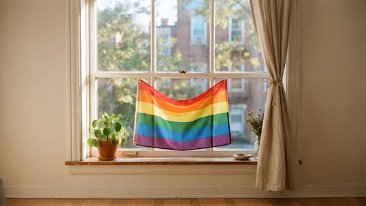 A six-color rainbow Pride flag hanging in a bright apartment window with a plant and curtains