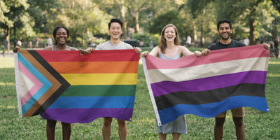 Four friends in a sunlit park holding two Pride Belongs 3x5 flags stretched between them, the Progress Pride flag and the genderfluid flag
