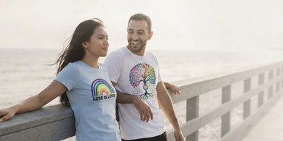 Two friends leaning on a seaside boardwalk rail in the late afternoon, wearing Pride Belongs Love is Love and Tree of Life rainbow tees